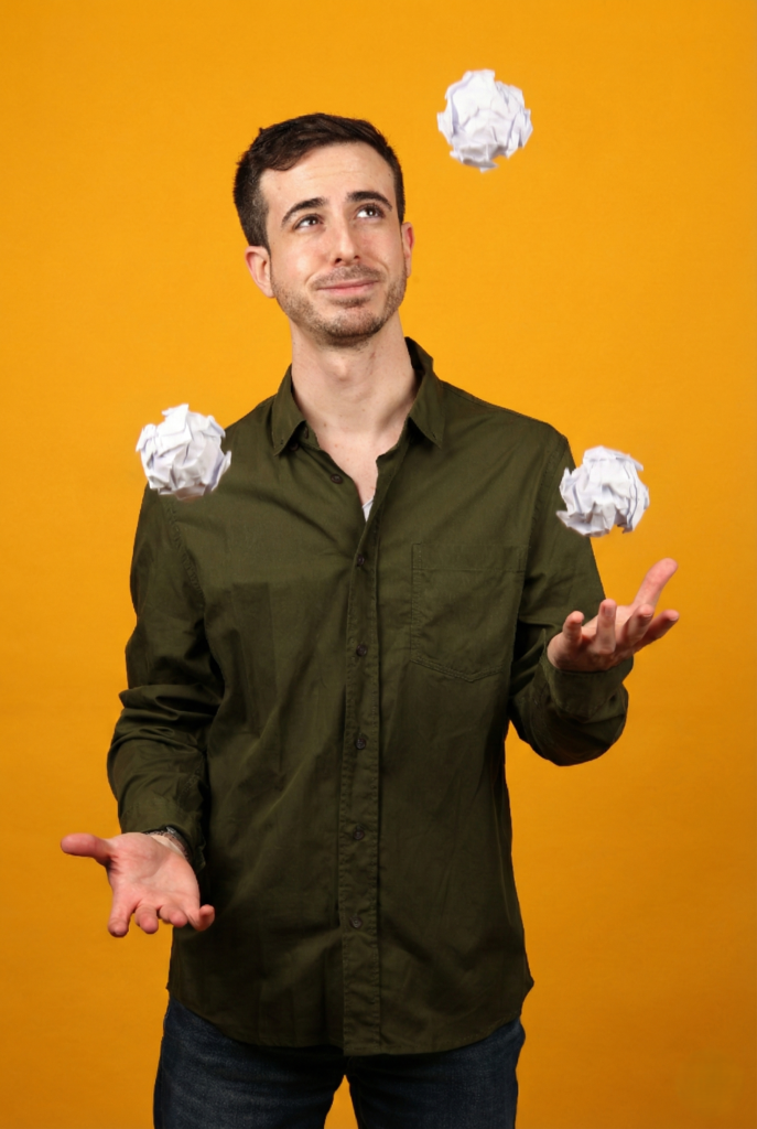 אורטל פלדמן זורק באוויר שלושה כדורי נייר לבנים מקומטים על רקע צהוב בוהק.