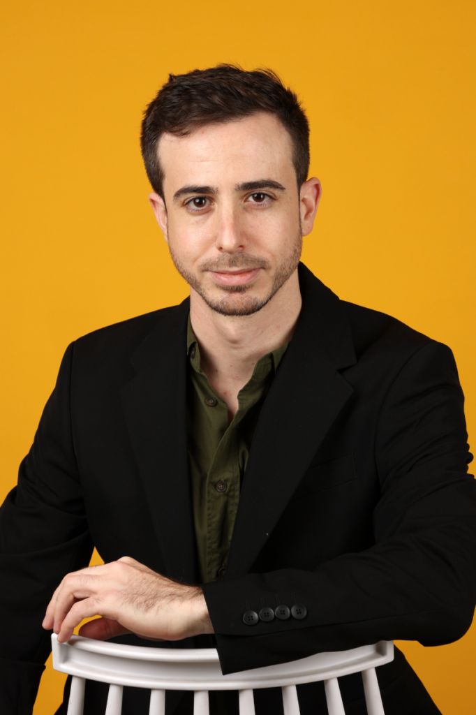 תמונת פורטרט של אורטל פלדמן, לבוש בז'קט שחור וחולצה ירוקה, על רקע צהוב בוהק.